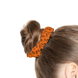 An orange fabric hair tie with a black pumpkin and spider pattern, shown securing a brown hair bun on a model's head against a white background.