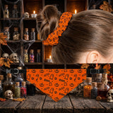 An orange Halloween-themed pet bandana with black printed cartoon ghosts, pumpkins, and hearts, shown hanging from clips on a rustic wooden table with a woman's head in the background wearing a matching scrunchie.