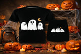 A black short-sleeve T-shirt and matching black shorts set with a white ghost graphic and a small orange crescent moon, displayed on a rustic Halloween-themed wooden stand with pumpkins, candles, spiderwebs, and autumn leaves.