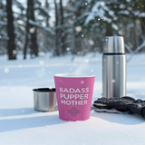 Pink cup with 'BADASS PUPPER MOTHER' text in a snowy outdoor setting with thermos and gloves.