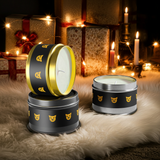 Decorative candles in black tins with gold accents on a soft surface with lights and presents in the background.