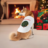 Dog wearing a Christmas-themed shirt in front of a fireplace and decorated tree.